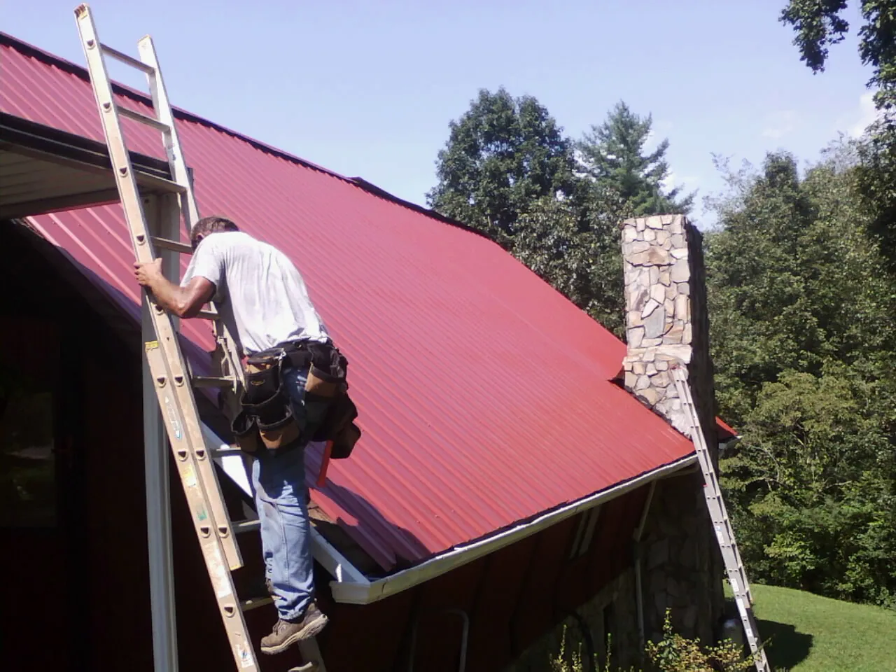 Roofer on a ladder beside a red metal roof and stone chimney — Soffit & Fascia Repair by Roofing Brothers in Seabeck